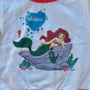 Op de afbeelding: Een wit sweatshirt met lange mouwen en een afbeelding van Ari&euml;l uit De Kleine Zeemeermin. Het shirt heeft een geribbelde opstaande kraag in turquoise en roze, en een roze geribbelde zoom. De print bevat een blauw hart, bubbels en Disney-logo.