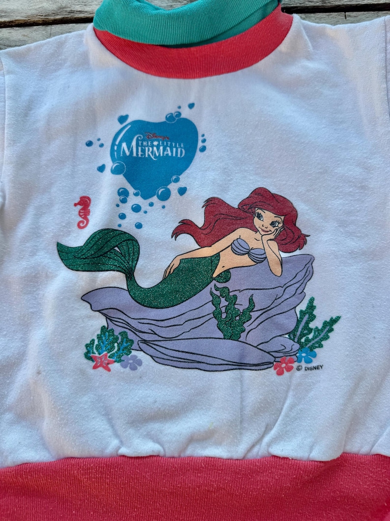 Op de afbeelding: Een wit sweatshirt met lange mouwen en een afbeelding van Ari&euml;l uit De Kleine Zeemeermin. Het shirt heeft een geribbelde opstaande kraag in turquoise en roze, en een roze geribbelde zoom. De print bevat een blauw hart, bubbels en Disney-logo.