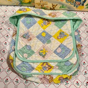 May include: A quilted baby carrier with a patchwork design featuring Cabbage Patch Kids characters, balloons, and pastel colors. The carrier has a mint green trim and a quilted pattern.
