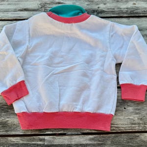 Op de afbeelding: Een wit sweatshirt met lange mouwen, voorzien van een felroze geribbelde kraag, boorden en zoom. Het kledingstuk heeft een contrasterende turquoise accentlaag zichtbaar bij de halslijn, wat zorgt voor een kleurrijk ontwerp aan de bovenzijde.
