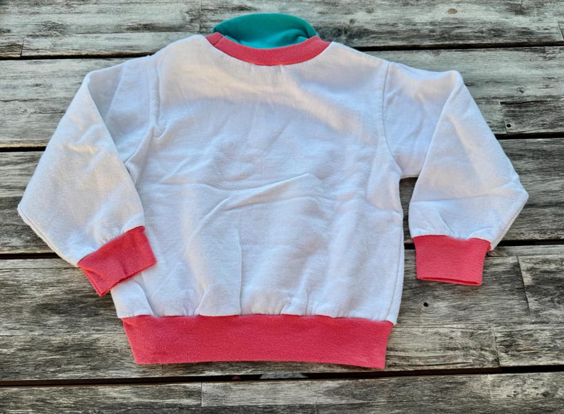Op de afbeelding: Een wit sweatshirt met lange mouwen, voorzien van een felroze geribbelde kraag, boorden en zoom. Het kledingstuk heeft een contrasterende turquoise accentlaag zichtbaar bij de halslijn, wat zorgt voor een kleurrijk ontwerp aan de bovenzijde.