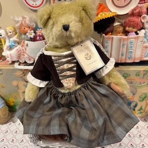 May include: A plush teddy bear with light brown fur wearing a dark brown bodice with a tan patterned lace-up front, white sleeves, and a grey and brown plaid skirt. A rectangular tag attached to the chest reads Outlander and Claire Randall.