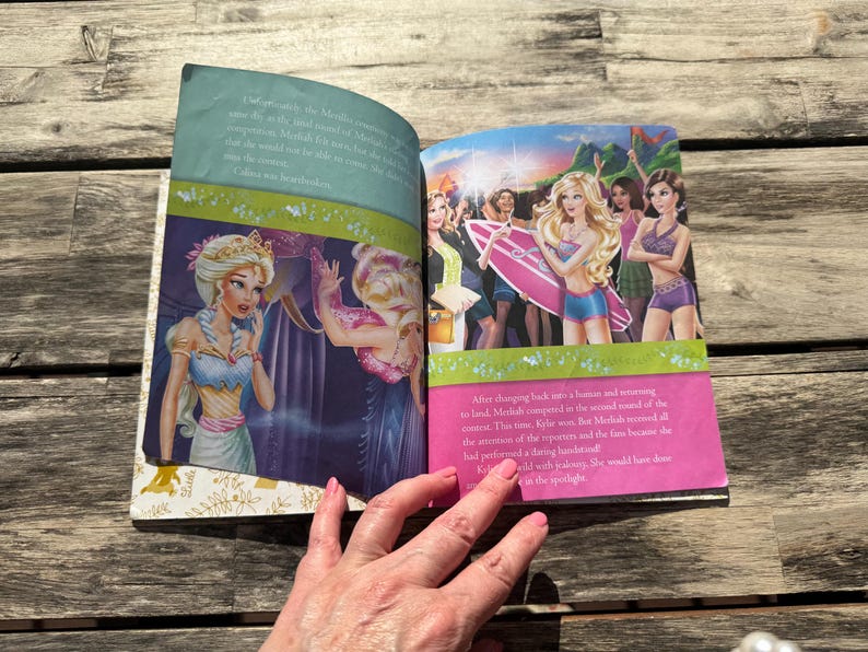 Pode incluir: Um livro infantil aberto em uma ilustra&ccedil;&atilde;o colorida. Uma p&aacute;gina mostra uma princesa com um vestido azul e branco. A outra p&aacute;gina mostra um grupo de meninas com uma prancha de surf. O texto &eacute; vis&iacute;vel em ambas as p&aacute;ginas.