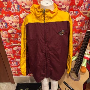 May include: A maroon and gold windbreaker jacket featuring a high collar, a full-length front zip, and long sleeves. The jacket displays a small embroidered mascot logo on the upper left chest area, showcasing a stylised character design.