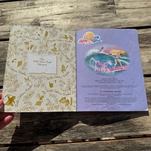 Pode incluir: Livro Little Golden Book aberto com uma ilustra&ccedil;&atilde;o da Barbie na p&aacute;gina direita. A p&aacute;gina esquerda tem um padr&atilde;o dourado e o texto "This Little Golden Book belongs to". O livro est&aacute; aberto sobre uma superf&iacute;cie de madeira.