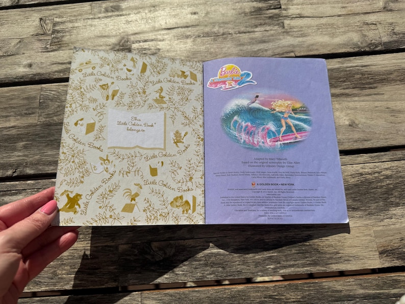 Pode incluir: Livro Little Golden Book aberto com uma ilustra&ccedil;&atilde;o da Barbie na p&aacute;gina direita. A p&aacute;gina esquerda tem um padr&atilde;o dourado e o texto "This Little Golden Book belongs to". O livro est&aacute; aberto sobre uma superf&iacute;cie de madeira.