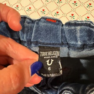 以下が含まれることがあります： 青いデニムジーンズのクローズアップ。タグには「TRUE RELIGION BRAND JEANS」とサイズ6のラベルが記載されています。ジーンズはパキスタン製です。