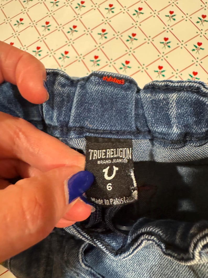 以下が含まれることがあります： 青いデニムジーンズのクローズアップ。タグには「TRUE RELIGION BRAND JEANS」とサイズ6のラベルが記載されています。ジーンズはパキスタン製です。