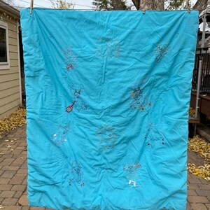 May include: A turquoise quilt with embroidered teddy bears and other whimsical figures. The quilt is hanging outside on a clothesline. The embroidery is in various colors, including red, yellow, and blue.