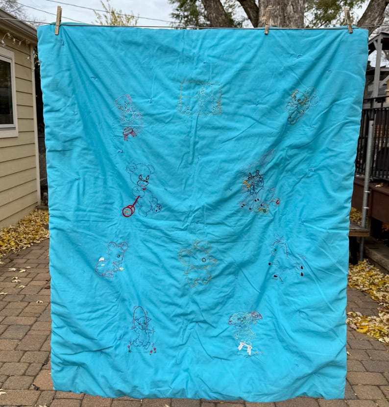 May include: A turquoise quilt with embroidered teddy bears and other whimsical figures. The quilt is hanging outside on a clothesline. The embroidery is in various colors, including red, yellow, and blue.