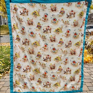 May include: A vintage quilt with a white background featuring a repeating pattern of whimsical figures, castles, and beach scenes in shades of red, yellow, and blue. The quilt is trimmed with a teal blue border.