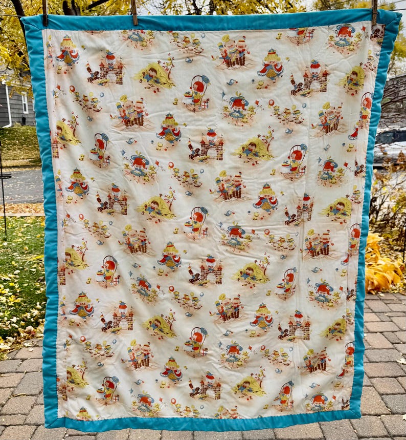 May include: A vintage quilt with a white background featuring a repeating pattern of whimsical figures, castles, and beach scenes in shades of red, yellow, and blue. The quilt is trimmed with a teal blue border.