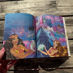 Pode incluir: Livro aberto com ilustra&ccedil;&otilde;es coloridas de sereias em uma cena subaqu&aacute;tica. As ilustra&ccedil;&otilde;es apresentam sereias com cabelos longos, segurando conchas e outros objetos. O fundo &eacute; uma mistura vibrante de azuis e roxos.