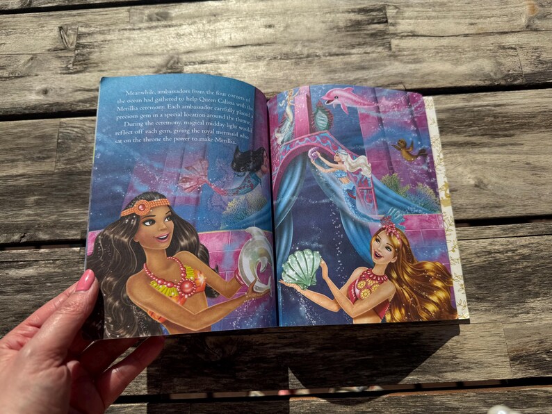 Pode incluir: Livro aberto com ilustra&ccedil;&otilde;es coloridas de sereias em uma cena subaqu&aacute;tica. As ilustra&ccedil;&otilde;es apresentam sereias com cabelos longos, segurando conchas e outros objetos. O fundo &eacute; uma mistura vibrante de azuis e roxos.
