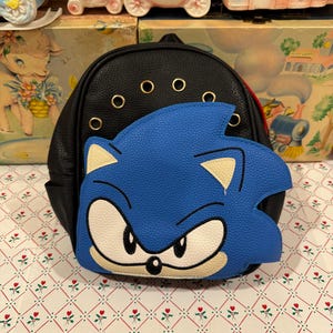 May include: A black faux leather mini backpack featuring a large, textured blue Sonic the Hedgehog character face applique on the front pocket. The bag includes gold-tone metal grommets along the top edge and a black zipper closure.