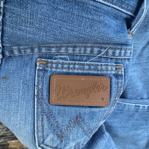 Puede incluir: Primer plano de unos vaqueros de mezclilla azul claro con un parche de cuero marr&oacute;n en el bolsillo trasero. El parche presenta el nombre de la marca "Wrangler" en cursiva. Los vaqueros tienen detalles de costura cl&aacute;sicos.