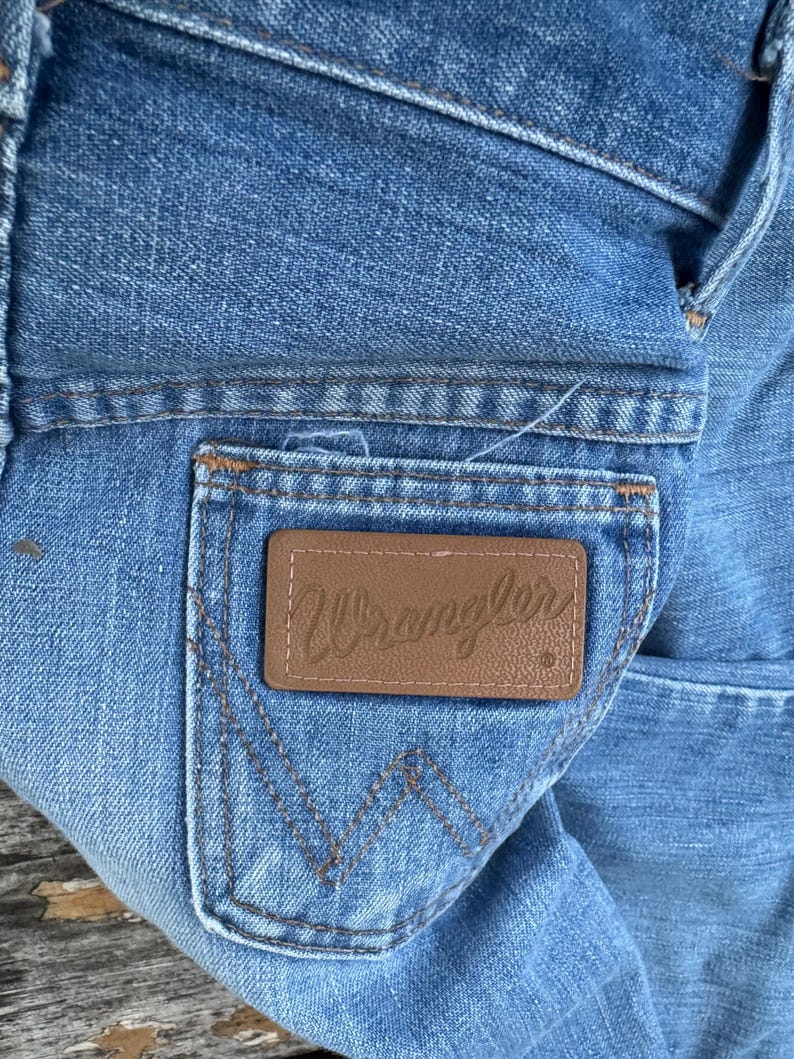 Puede incluir: Primer plano de unos vaqueros de mezclilla azul claro con un parche de cuero marr&oacute;n en el bolsillo trasero. El parche presenta el nombre de la marca "Wrangler" en cursiva. Los vaqueros tienen detalles de costura cl&aacute;sicos.