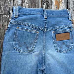 Puede incluir: Primer plano de la parte trasera de unos vaqueros de mezclilla azul claro. Incluye dos bolsillos traseros con costuras decorativas y un parche de cuero marr&oacute;n con la marca "Wrangler".