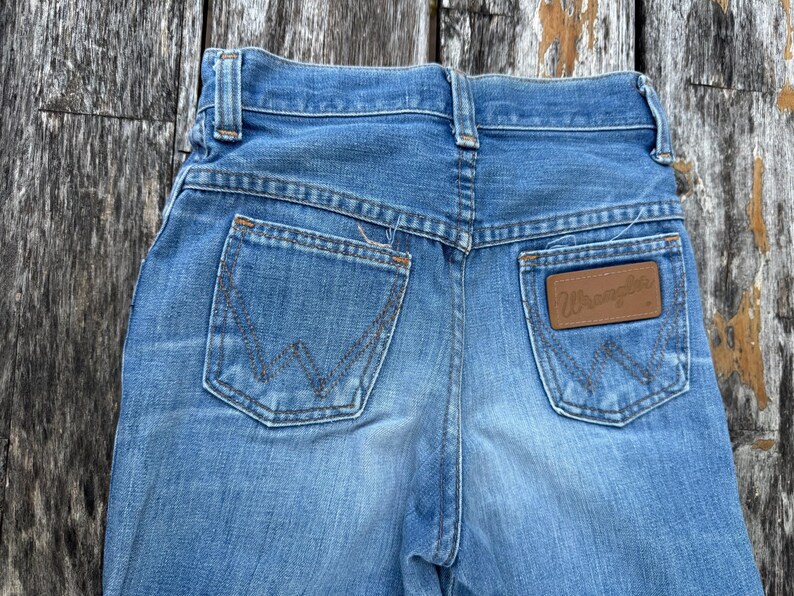 Puede incluir: Primer plano de la parte trasera de unos vaqueros de mezclilla azul claro. Incluye dos bolsillos traseros con costuras decorativas y un parche de cuero marr&oacute;n con la marca "Wrangler".