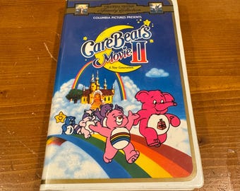 May include: A vintage VHS tape for "Care Bears Movie II" featuring a colorful design. The cover displays the title in a playful font, a rainbow, and cartoon Care Bears characters. The tape is from the Columbia Tristar Family Collection.