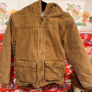 Puede incluir: Chaqueta con capucha Carhartt marr&oacute;n con cierre de cremallera completo. La chaqueta tiene dos bolsillos en el pecho y dos bolsillos inferiores. El logotipo de Carhartt es visible en el bolsillo derecho. La chaqueta est&aacute; hecha de tela resistente.