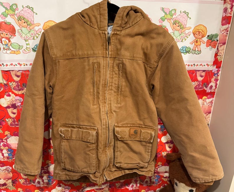 Puede incluir: Chaqueta con capucha Carhartt marr&oacute;n con cierre de cremallera completo. La chaqueta tiene dos bolsillos en el pecho y dos bolsillos inferiores. El logotipo de Carhartt es visible en el bolsillo derecho. La chaqueta est&aacute; hecha de tela resistente.