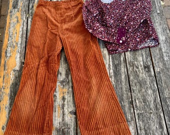 Op de afbeelding: Een roestkleurige corduroy broek met wijde pijpen en een donkerpaarse blouse met lange mouwen en bloemenprint, voorzien van een witte kanten kraag en een knoopsluiting met één zichtbare witte knoop op de manchet.