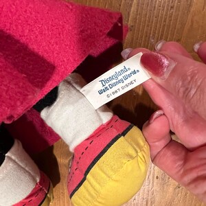 May include: A plush Mickey Mouse doll wearing a red hooded cloak. The doll has yellow shoes with black stitching. A white tag attached to the doll reads "Disneyland Walt Disney World &copy;1987 DISNEY".