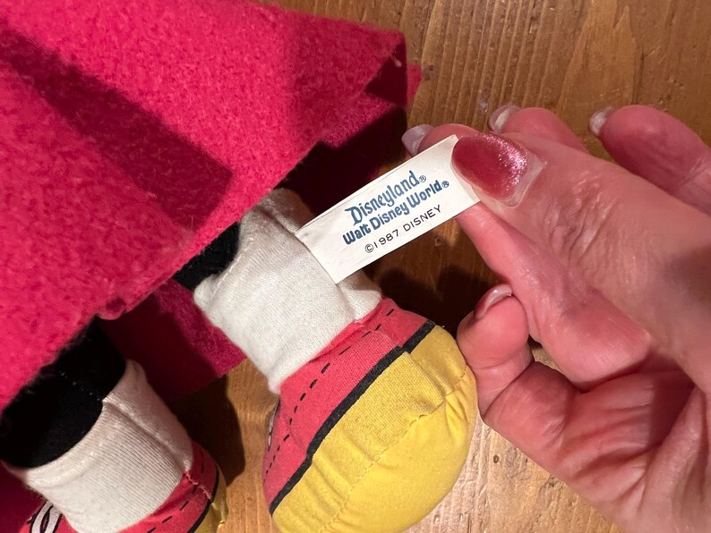 May include: A plush Mickey Mouse doll wearing a red hooded cloak. The doll has yellow shoes with black stitching. A white tag attached to the doll reads "Disneyland Walt Disney World &copy;1987 DISNEY".