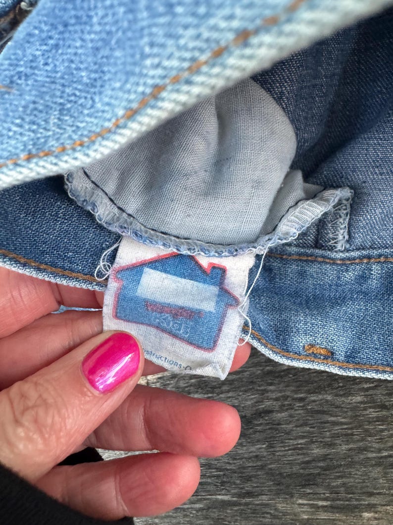 Puede incluir: Primer plano de una prenda de mezclilla azul claro, posiblemente unos vaqueros o una chaqueta, con una etiqueta visible. La etiqueta presenta un gr&aacute;fico de casa en azul y blanco, con texto parcialmente visible. Tambi&eacute;n se ven las costuras y la textura de la tela.