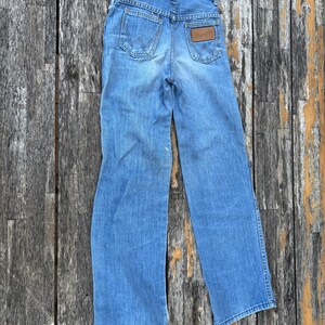 Puede incluir: Vaqueros de mezclilla azul claro con el logotipo de Wrangler en los bolsillos traseros. Los vaqueros tienen un dise&ntilde;o cl&aacute;sico de cinco bolsillos y un parche de cuero marr&oacute;n. Los vaqueros est&aacute;n planos sobre una superficie de madera.
