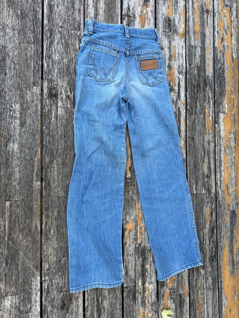 Puede incluir: Vaqueros de mezclilla azul claro con el logotipo de Wrangler en los bolsillos traseros. Los vaqueros tienen un dise&ntilde;o cl&aacute;sico de cinco bolsillos y un parche de cuero marr&oacute;n. Los vaqueros est&aacute;n planos sobre una superficie de madera.