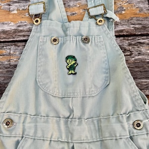 Puede incluir: Peto vaquero azul claro con tirantes ajustables y herrajes metálicos. El bolsillo del peto tiene un parche verde con un personaje de dibujos animados. Cierre con botones, para niños.