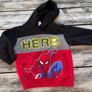 May include: A long-sleeve hooded sweatshirt featuring a color-blocked design in black, gray, and red. The gray chest panel displays the yellow text HERO with a spiderweb icon, while the red lower section features a graphic of Spider-Man.