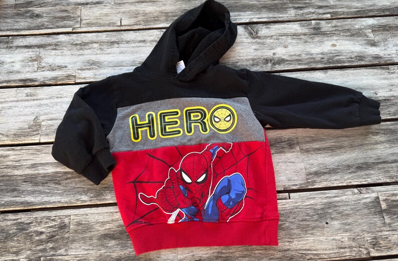 May include: A long-sleeve hooded sweatshirt featuring a color-blocked design in black, gray, and red. The gray chest panel displays the yellow text HERO with a spiderweb icon, while the red lower section features a graphic of Spider-Man.