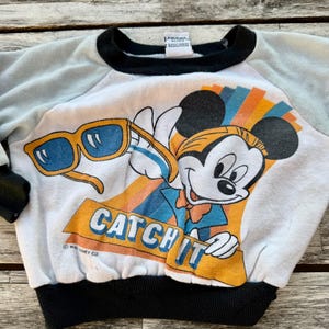 May include: A white sweatshirt with light blue sleeves and black trim at the collar and waist. The front features a graphic of Mickey Mouse holding yellow sunglasses with blue lenses, above the orange text CATCH IT and Walt Disney Co.
