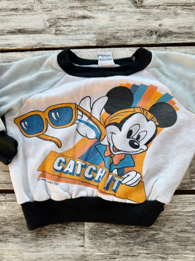 May include: A white sweatshirt with light blue sleeves and black trim at the collar and waist. The front features a graphic of Mickey Mouse holding yellow sunglasses with blue lenses, above the orange text CATCH IT and Walt Disney Co.