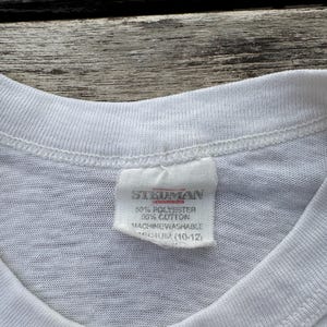 May include: A white t-shirt featuring a sewn-in fabric label at the inner neckline. The label displays the brand name Stedman, material composition of 50% polyester and 50% cotton, machine washable instructions, and a size medium 10-12 label.