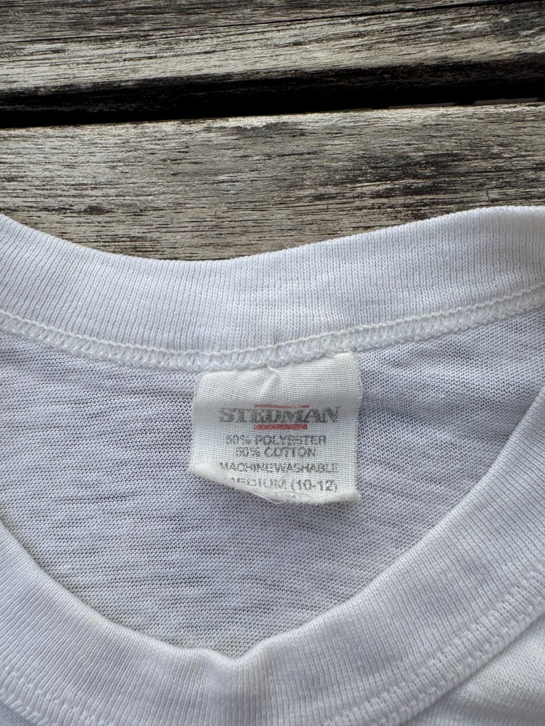 May include: A white t-shirt featuring a sewn-in fabric label at the inner neckline. The label displays the brand name Stedman, material composition of 50% polyester and 50% cotton, machine washable instructions, and a size medium 10-12 label.