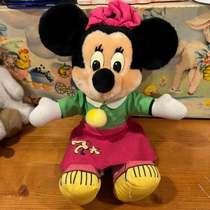 May include: A plush Minnie Mouse doll wearing a green dress, pink skirt, and yellow shoes. She has a pink bow on her head and a yellow pom-pom on her dress.