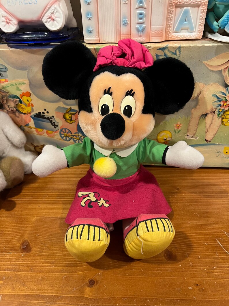 May include: A plush Minnie Mouse doll wearing a green dress, pink skirt, and yellow shoes. She has a pink bow on her head and a yellow pom-pom on her dress.