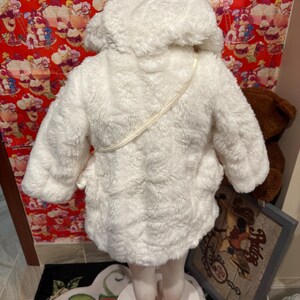 May include: A white, fluffy hooded coat, likely for a child, with a soft, textured appearance. The coat has a hood and a strap across the back. The coat is displayed on a mannequin.