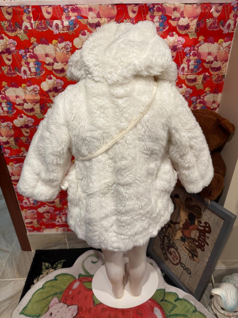 May include: A white, fluffy hooded coat, likely for a child, with a soft, textured appearance. The coat has a hood and a strap across the back. The coat is displayed on a mannequin.