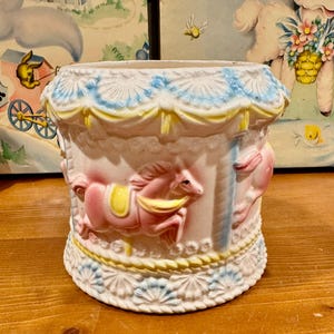 May include: A vintage ceramic carousel planter in a soft pastel color palette. The planter features pink carousel horses with yellow saddles, blue and yellow decorative trim, and a scalloped base. The overall design is reminiscent of a classic carousel.