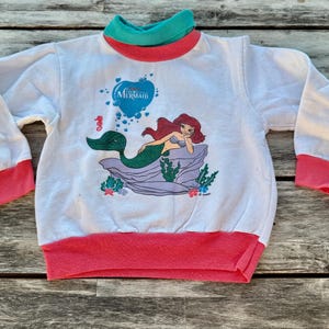 Op de afbeelding: Een wit sweatshirt met lange mouwen en een afbeelding van Ari&euml;l de kleine zeemeermin die op een rots rust. Het kledingstuk heeft een blauwgroene opstaande kraag, roze mouwboorden en een roze tailleband. De afbeelding bevat een blauw hart met tekst.