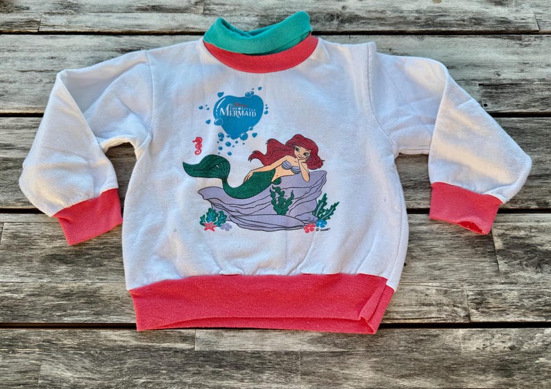 Op de afbeelding: Een wit sweatshirt met lange mouwen en een afbeelding van Ari&euml;l de kleine zeemeermin die op een rots rust. Het kledingstuk heeft een blauwgroene opstaande kraag, roze mouwboorden en een roze tailleband. De afbeelding bevat een blauw hart met tekst.