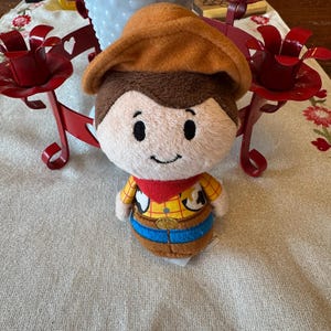 May include: A soft toy depicting a cartoon cowboy character, with a brown hat, brown hair, and a smiling face. The toy is dressed in a yellow shirt with a brown vest, blue trousers, and a red bandana. The toy is on a cream-coloured surface.
