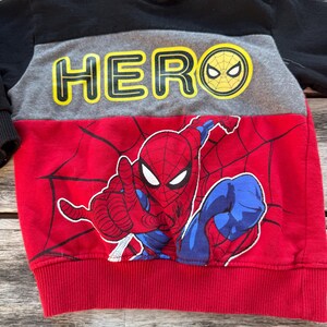 May include: A color-blocked hooded sweatshirt featuring a black hood and sleeves, a gray chest panel with the yellow text HERO, and a red lower section with a graphic print of Spider-Man and black spiderweb patterns on the front.