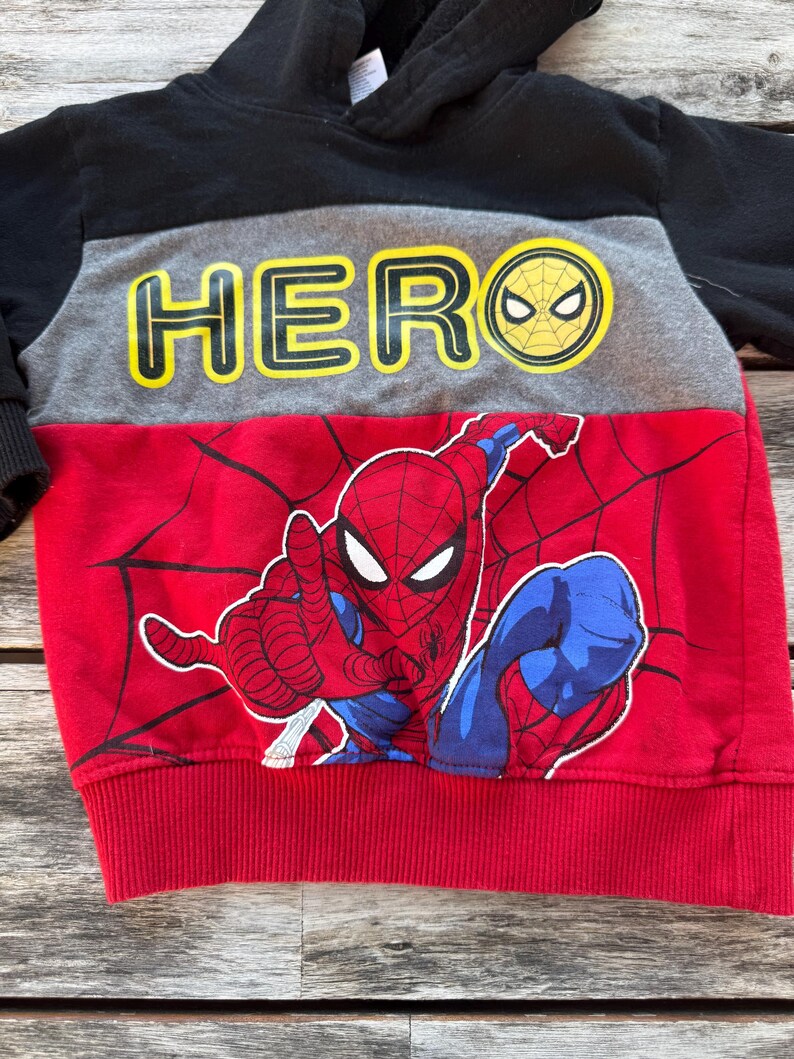 May include: A color-blocked hooded sweatshirt featuring a black hood and sleeves, a gray chest panel with the yellow text HERO, and a red lower section with a graphic print of Spider-Man and black spiderweb patterns on the front.