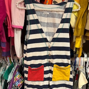May include: A sleeveless striped dress with a deep V-neckline and two pockets. The pockets are red and yellow with green trim. The dress has a button closure at the neckline and is made of a soft, comfortable fabric.